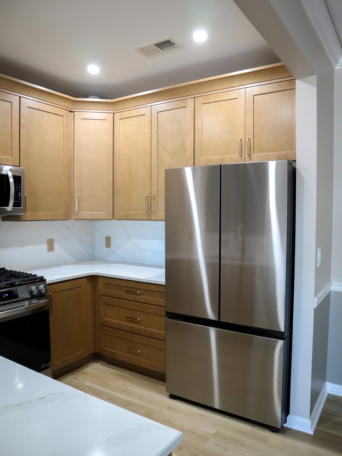 The same kitchen after the remodel, wood-tone shaker cabinets, white quartz and stainless appliances